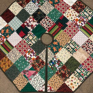 May include: A patchwork Christmas tree skirt with a variety of festive patterns. The skirt is octagonal, featuring squares of green, red, and beige fabrics with holiday motifs like gingerbread men, stars, and presents. A dark brown rope trim edges the skirt.