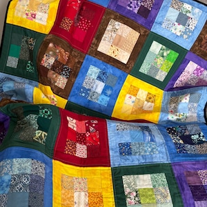 May include: A handmade quilt with a patchwork design. The quilt features squares of various colors, including red, yellow, blue, green, and purple. Each square is bordered by a darker color, creating a framed effect. The quilt is folded over a surface.