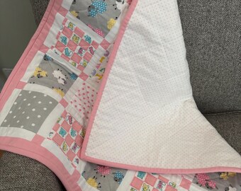 Puede incluir: Una colcha de bebé hecha a mano con un diseño de patchwork. La colcha presenta cuadrados de gris con lunares blancos, rosa y tela blanca. Algunos cuadrados tienen diseños de ovejas y animales bordados. La colcha está ribeteada con un ribete rosa.