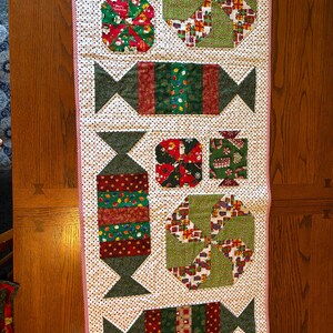 May include: A festive Christmas table runner featuring candy and gift box designs. The runner is made of fabric with a white background and small black polka dots. The candy shapes are red, green, and brown. The edges are trimmed with a red and white striped fabric.