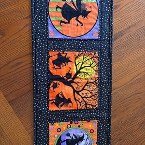 May include: A rectangular quilted table runner featuring a black fabric border with a yellow star pattern. The runner contains three square panels depicting black witch silhouettes, orange and purple backgrounds, and spooky tree motifs.