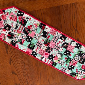 May include: A quilted table runner with a patchwork design. The runner features a red border and a pattern of squares in black, white, pink, and mint green. The squares are decorated with cherries, polka dots, and floral prints.