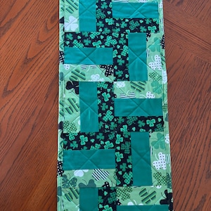 Pode incluir: Um caminho de mesa acolchoado verde e preto com tema do Dia de São Patrício. O caminho de mesa apresenta um design de patchwork com trevos e padrões geométricos. O caminho de mesa tem aproximadamente 30 cm de largura e 91 cm de comprimento.