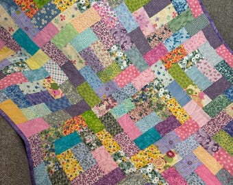 May include: A colorful patchwork quilt with a variety of fabric patterns and colors, including pink, purple, green, yellow, and blue. The quilt has a purple border and is likely made of cotton. The design is a mix of floral, polka dot, and geometric patterns.