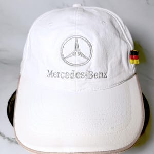May include: A white baseball cap featuring the embroidered Mercedes-Benz logo and text in silver thread on the front panel. A small fabric tag with the black, red, and gold colors of the German flag is attached to the side of the cap brim.