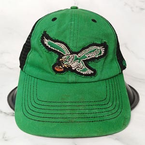 Könnte beinhalten: Grüne Baseballkappe mit schwarzem Mesh-Rücken und gebogenem Schirm. Die Kappe hat einen aufgenähten Aufnäher eines fliegenden Adlers in Weiß, Grün und Schwarz. Die Kappe steht auf einem schwarzen Ständer vor einem weißen Marmorhintergrund.