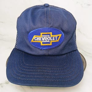 Könnte beinhalten: Marineblaue Trucker-Cap mit dem Chevrolet-Logo in Gelb und Weiß auf einem blauen ovalen Aufnäher. Die Kappe hat einen Mesh-Rücken und einen gebogenen Schirm. Hergestellt aus strapazierfähigem Stoff.