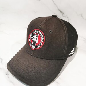 Puede incluir: Gorra de b&eacute;isbol negra con el logotipo del North Mississauga Soccer Club, un emblema circular rojo y blanco. El logotipo de Adidas es visible en el lateral. La gorra tiene una visera curva.