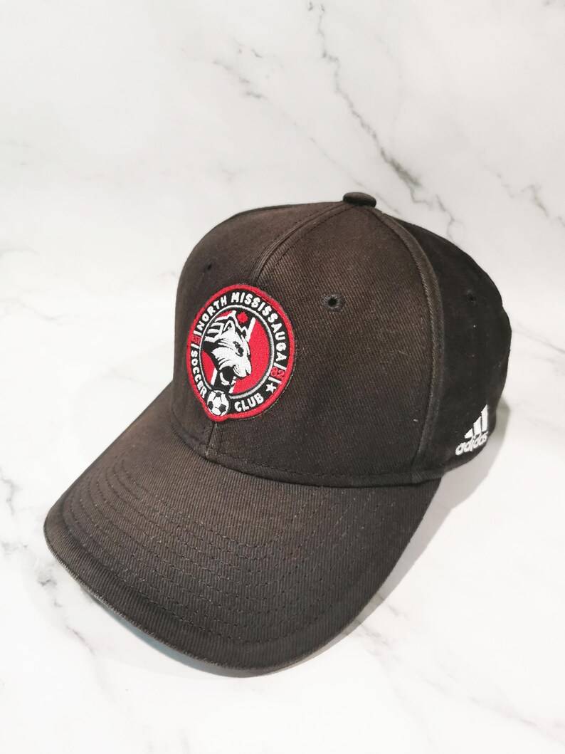 Puede incluir: Gorra de b&eacute;isbol negra con el logotipo del North Mississauga Soccer Club, un emblema circular rojo y blanco. El logotipo de Adidas es visible en el lateral. La gorra tiene una visera curva.