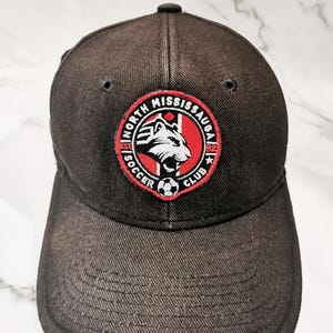 Puede incluir: Gorra de b&eacute;isbol gris oscuro con un parche circular con el texto "NORTH MISSISSAUGA SOCCER CLUB" y una cabeza de lobo estilizada. El parche es rojo, blanco y negro. La gorra tiene una visera curva y un bot&oacute;n en la parte superior.