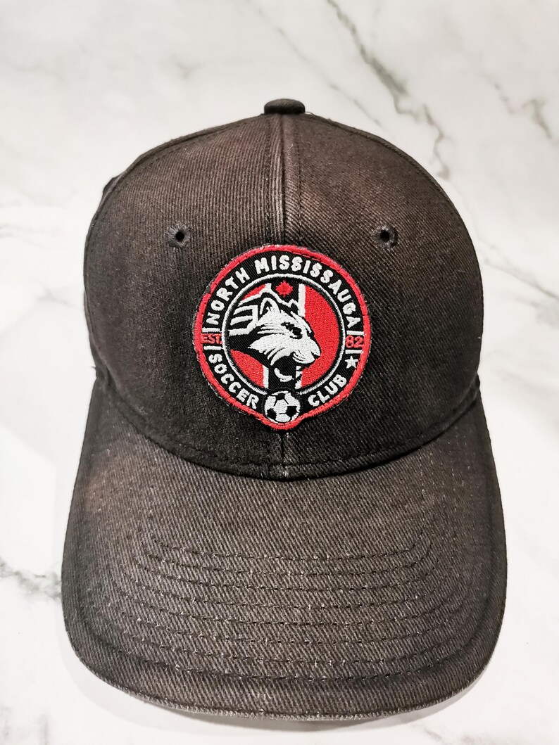 Puede incluir: Gorra de b&eacute;isbol gris oscuro con un parche circular con el texto "NORTH MISSISSAUGA SOCCER CLUB" y una cabeza de lobo estilizada. El parche es rojo, blanco y negro. La gorra tiene una visera curva y un bot&oacute;n en la parte superior.