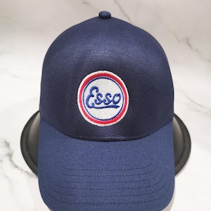 Könnte beinhalten: Marineblaue Baseballkappe mit gebogenem Schirm. Auf der Kappe befindet sich ein kreisförmiges Emblem mit dem Wort "Esso" in weißer Schrift, umrandet von Rot und Blau. Hergestellt aus strukturiertem Stoff.