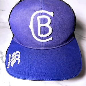 Peut inclure: Une casquette de baseball bleue avec un dos en maille noire. Le panneau avant présente une grande lettre B brodée en blanc. La visière affiche un logo Canterbury brodé en blanc et le texte canterbury orienté verticalement sur le côté gauche.