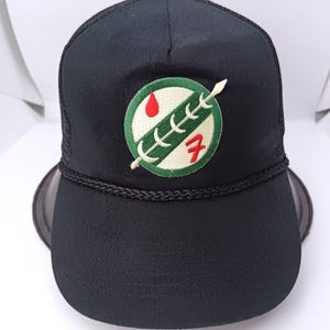 Op de afbeelding: Zwarte truckerpet met een mesh achterkant en een gebogen klep. De voorkant heeft een cirkelvormige patch met een groen en wit ontwerp, een rode druppel en het nummer 7. Een zwart touwdetail accentueert de klep.