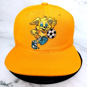 Puede incluir: Gorra de béisbol naranja brillante con visera negra. La gorra presenta un perro de dibujos animados bordado jugando al fútbol. El perro es amarillo con botas de fútbol azules y un balón de fútbol blanco y negro. La gorra es de estilo visera plana.