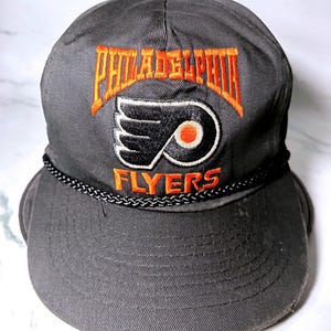 Peut inclure: Une casquette de baseball gris anthracite avec le logo brodé des Philadelphia Flyers en noir et orange. Le mot Philadelphia est arqué au-dessus du logo en orange, avec le mot Flyers en dessous en orange. Un cordon tressé noir orne la visière.