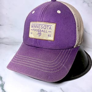 Pode incluir: Um boné de beisebol roxo com parte traseira de rede bege e um emblema retangular na frente. O emblema apresenta o texto MINNESOTA FOOTBALL, os números 19 e 61 e um pequeno logótipo viking roxo. O boné tem costuras brancas na aba.