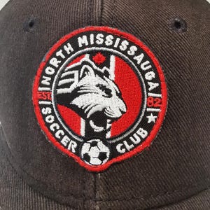 Puede incluir: Gorra de b&eacute;isbol marr&oacute;n oscuro con un parche circular. El parche muestra el texto "North Mississauga Soccer Club", una cabeza de tigre blanca y negra, un bal&oacute;n de f&uacute;tbol y un borde rojo.