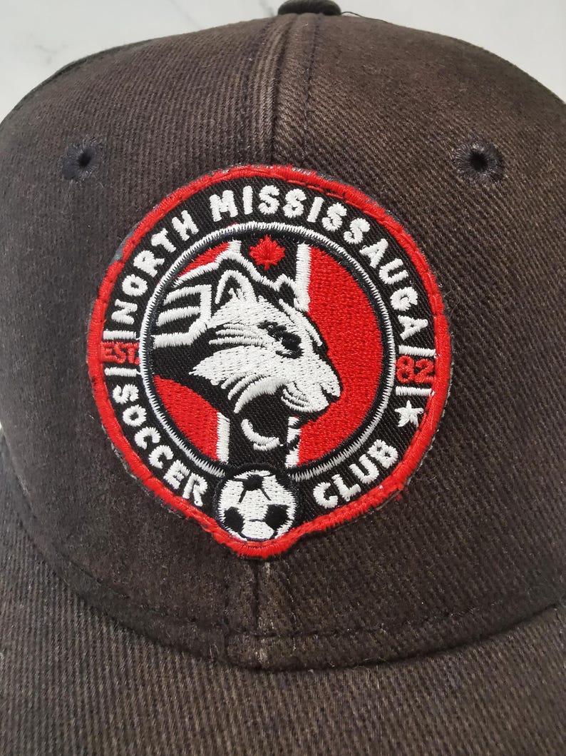 Puede incluir: Gorra de b&eacute;isbol marr&oacute;n oscuro con un parche circular. El parche muestra el texto "North Mississauga Soccer Club", una cabeza de tigre blanca y negra, un bal&oacute;n de f&uacute;tbol y un borde rojo.