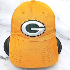 Puede incluir: Gorra de béisbol de color amarillo mostaza con visera curva. La gorra presenta un parche ovalado verde y blanco con un logotipo "G" estilizado. La gorra está hecha de una tela duradera y tiene un diseño clásico de seis paneles.