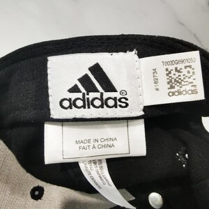 Puede incluir: Gorra de b&eacute;isbol negra con el logotipo de Adidas y texto. El logotipo de Adidas es blanco y negro. La etiqueta dice "MADE IN CHINA FAIT A CHINA". La gorra tiene una etiqueta blanca con texto y un c&oacute;digo QR.