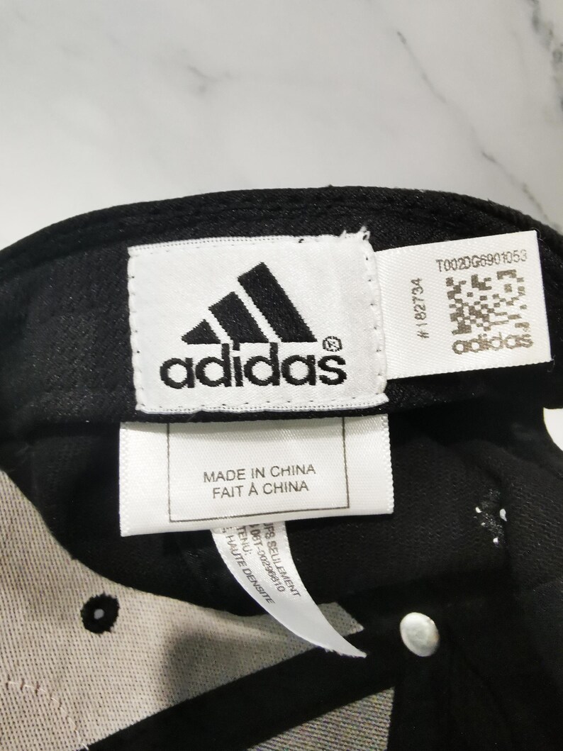 Puede incluir: Gorra de b&eacute;isbol negra con el logotipo de Adidas y texto. El logotipo de Adidas es blanco y negro. La etiqueta dice "MADE IN CHINA FAIT A CHINA". La gorra tiene una etiqueta blanca con texto y un c&oacute;digo QR.