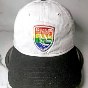 May include: A white baseball cap with a black brim featuring an embroidered shield-shaped patch on the front. The patch displays a rainbow-colored background with the text QUAKES SAN JOSE 1974 and a soccer ball graphic in the center.