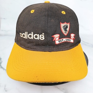 May include: A black and yellow baseball cap with the Adidas logo embroidered in white. The cap has a yellow peak and a crest with a bird and the text "EST. 1892".
