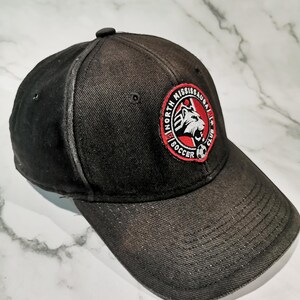 Puede incluir: Gorra de b&eacute;isbol negra con visera curva. La gorra presenta un parche circular con las palabras "North Mississauga Soccer Club" y un logotipo de cabeza de le&oacute;n en rojo, blanco y negro.