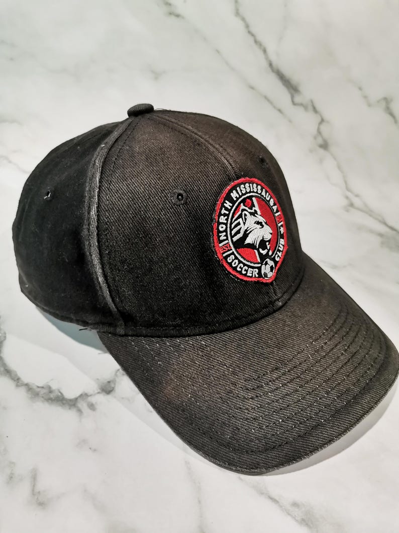 Puede incluir: Gorra de b&eacute;isbol negra con visera curva. La gorra presenta un parche circular con las palabras "North Mississauga Soccer Club" y un logotipo de cabeza de le&oacute;n en rojo, blanco y negro.