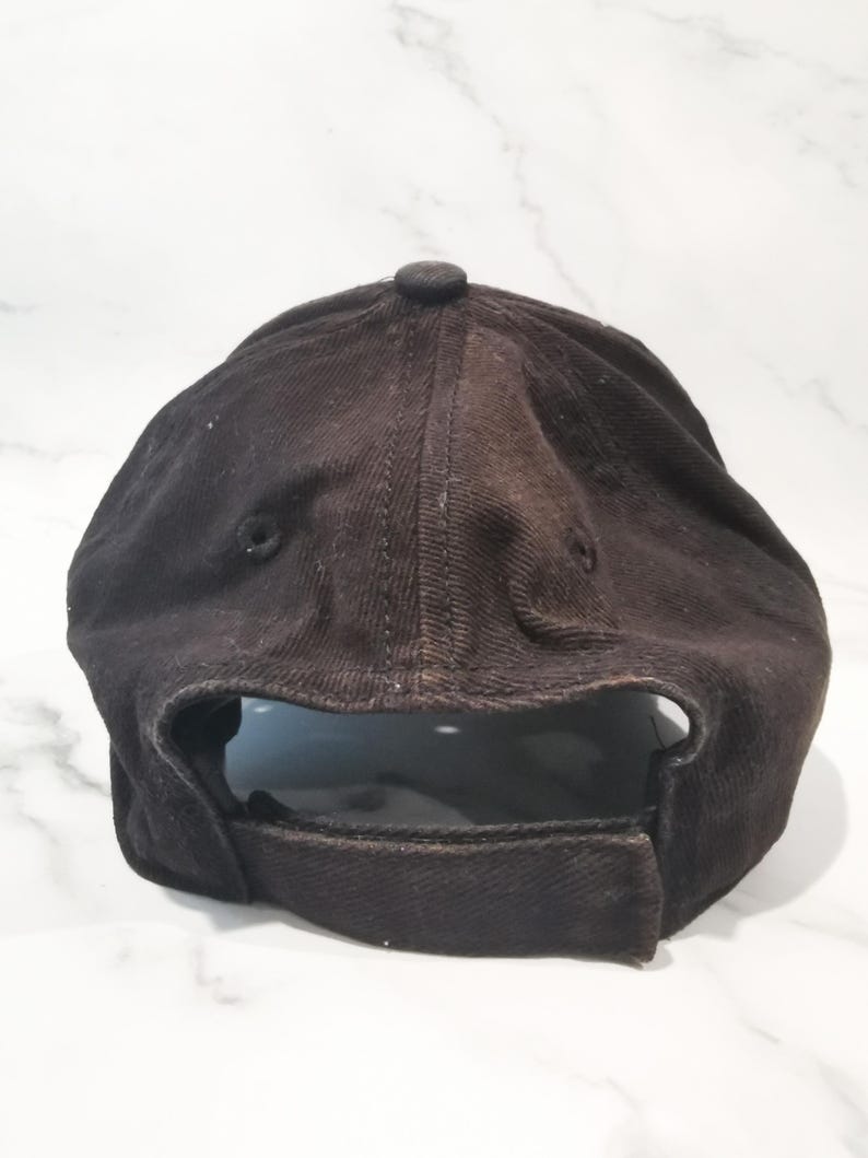 Puede incluir: Gorra de b&eacute;isbol negra con visera curva y correa ajustable. La gorra est&aacute; hecha de una tela texturizada y tiene un bot&oacute;n en la parte superior. La parte trasera de la gorra es visible, mostrando la correa ajustable.