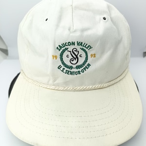 Op de afbeelding: Crèmekleurige baseballpet met een gebogen klep en een wit touwdetail. De pet heeft een geborduurd embleem met de tekst "Saucon Valley U.S. Senior Open" in groen en wit, met de jaren "79" en "92".