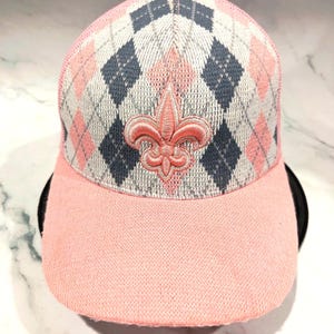 May include: A pink baseball cap with a knit brim and a mesh back. The front panel features a gray, pink, and navy blue argyle pattern and a pink fleur-de-lis emblem.