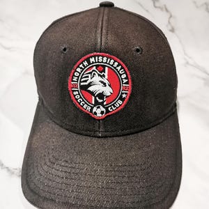 Puede incluir: Gorra de b&eacute;isbol negra con el logotipo del North Mississauga Soccer Club. El logotipo es un emblema circular con una cabeza de lobo blanca, el texto "North Mississauga Soccer Club" y el a&ntilde;o "82".