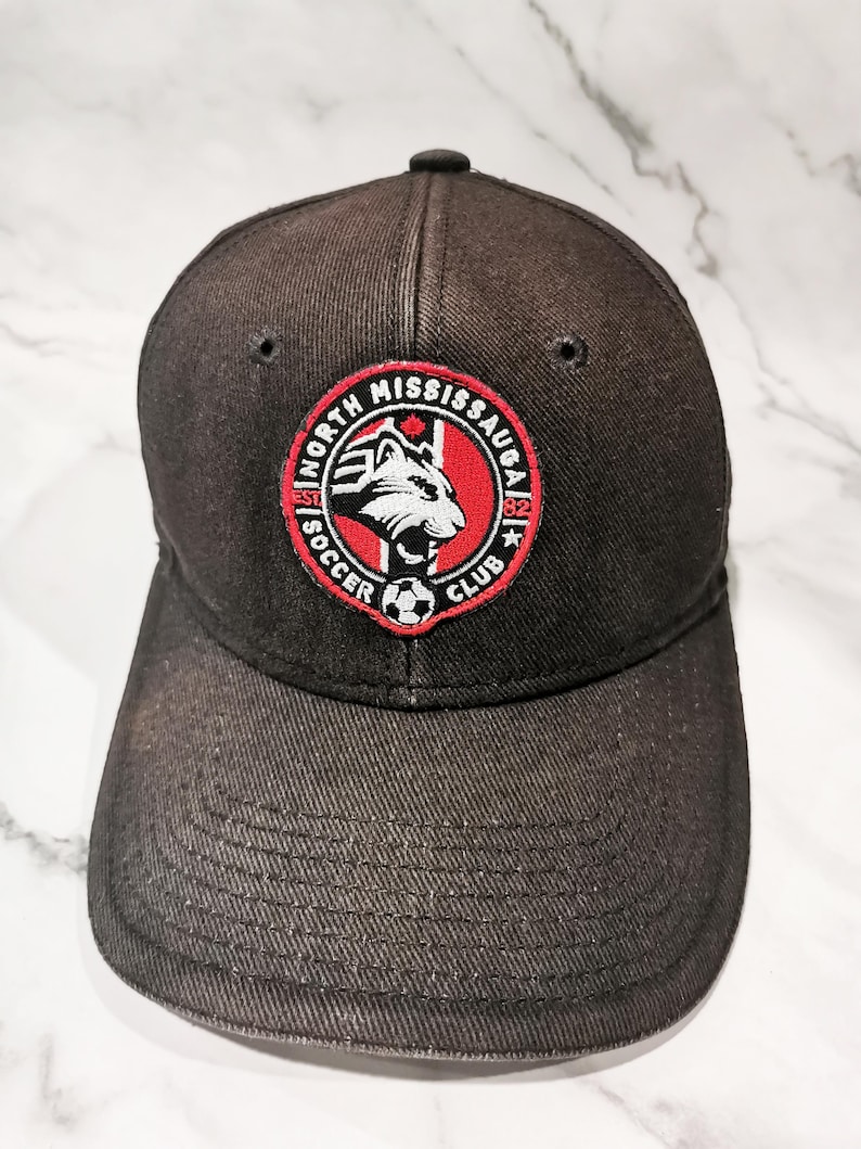 Puede incluir: Gorra de b&eacute;isbol negra con el logotipo del North Mississauga Soccer Club. El logotipo es un emblema circular con una cabeza de lobo blanca, el texto "North Mississauga Soccer Club" y el a&ntilde;o "82".