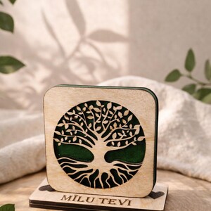 Puede incluir: Un posavasos de madera con un diseño de árbol de la vida. El posavasos cuadrado tiene un interior verde y un diseño de árbol cortado con láser. La base tiene la inscripción "MILU TEVI". El posavasos se muestra sobre una superficie de madera.