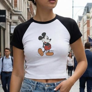 May include: White crop top with black sleeves featuring a Mickey Mouse graphic. The shirt is a baseball style tee. The image also shows a person wearing blue jeans. The shirt has the words "White" and "Black" on the bottom right.
