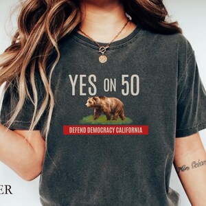 May include: Dark grey t-shirt with the text "YES ON 50" above a brown bear graphic on green grass. Below the bear is a red banner that reads "DEFEND DEMOCRACY CALIFORNIA".