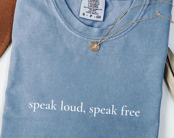 May include: A blue jean colored t-shirt with the words "speak loud, speak free" printed in white. A gold necklace with a pendant rests on the shirt. The shirt is folded and has a Comfort Colors tag.