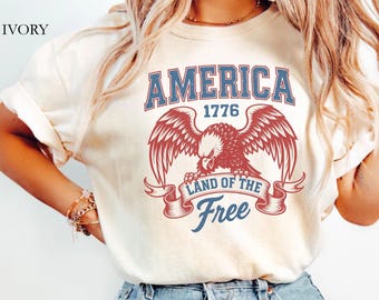May include: Ivory-colored t-shirt featuring a patriotic design. The graphic includes the word "AMERICA" above an eagle with outstretched wings, the year "1776", and the phrase "LAND OF THE FREE" on a banner. The shirt is paired with blue jeans.