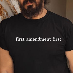 May include: Black crew neck t-shirt with the white text "first amendment first". The shirt is made of a soft material and has short sleeves. The text is centered on the front of the shirt.