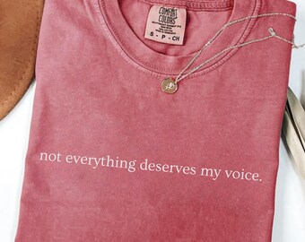 May include: Crimson-colored t-shirt with the text "not everything deserves my voice." A silver necklace with a pendant rests on the shirt. The shirt is folded, and a brown hat is visible in the background.