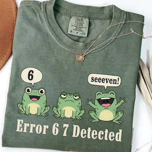 May include: A moss green t-shirt with a graphic of three cartoon frogs and the text "Error 6 7 Detected." The frogs have speech bubbles with the numbers "6" and the word "seeeven!" A silver necklace with a pendant is on the shirt.