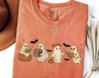 May include: Coral-colored long-sleeve shirt featuring a graphic of four cartoon ghosts reading books, with pumpkins, bats, and stars. The shirt has a crew neck and a Comfort Colors tag. The word "YAM" is printed at the bottom left.