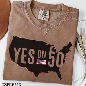 May include: Espresso-colored t-shirt with the text "YES ON 50" in a dark brown font, set against a black outline of the United States. The American flag is featured below the text. A silver necklace with a pendant is draped across the shirt.