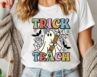 May include: White t-shirt with a colorful graphic that says "Trick or Teach". The design features a ghost holding a pencil, surrounded by bats, stars, and a spiderweb. A fun, festive design for Halloween or teachers.