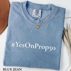 May include: A blue jean colored t-shirt with the text "#YesOnProp50" printed in white. A gold necklace with a pendant is draped over the shirt. The shirt is folded and has a tag that says "Comfort Colors".