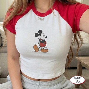May include: White crop top with red short sleeves featuring a classic Mickey Mouse graphic. The Disney-branded shirt has a crew neck and a fitted silhouette. The shirt is made of a soft, comfortable material.