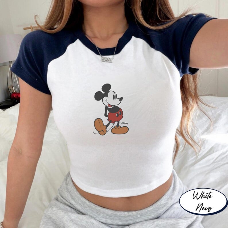 May include: White and navy blue raglan crop top featuring a Mickey Mouse graphic. The shirt has a white body with navy blue sleeves and neckline. The Mickey Mouse graphic is in the center of the shirt. The Disney logo is printed below the graphic.