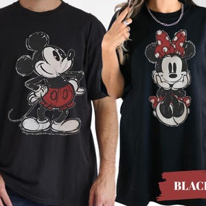 May include: Black t-shirts featuring Mickey Mouse and Minnie Mouse. Mickey is in a red shorts and Minnie is wearing a red bow. The graphic design has a distressed, vintage look. The word "BLACK" is in a red rectangle.
